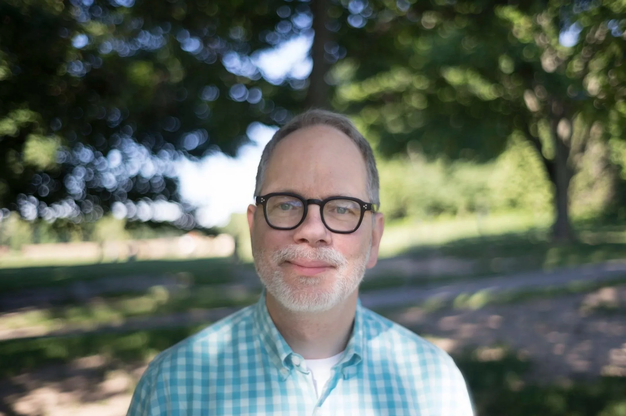 Close-up of a smiling middle-aged man with gray hair and beard, wearing glasses and a blue checkered shirt, outdoors in a park with green trees and sunlight in the background.