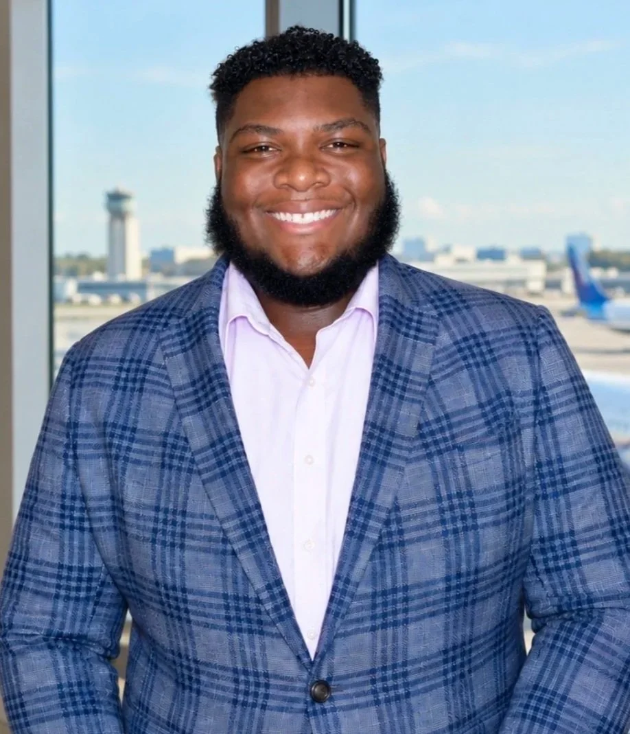 A smiling man with curly black hair and a beard, wearing a blue plaid blazer and a light pink shirt, standing in front of a window with an airport tarmac and a plane in the background.