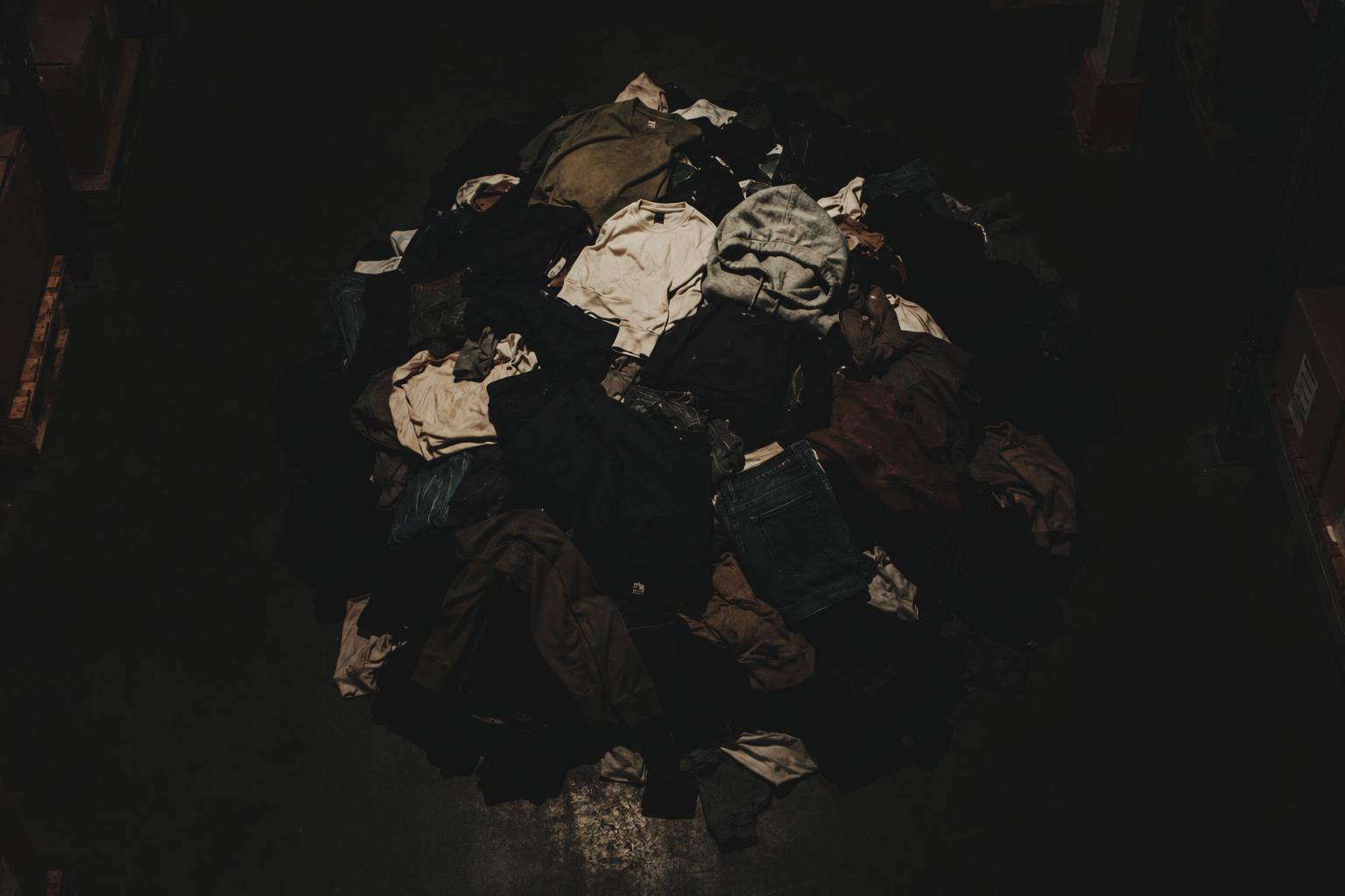 Pile of mixed clothes, including shirts, pants, and a hoodie, on the dark floor of a warehouse or storage area.