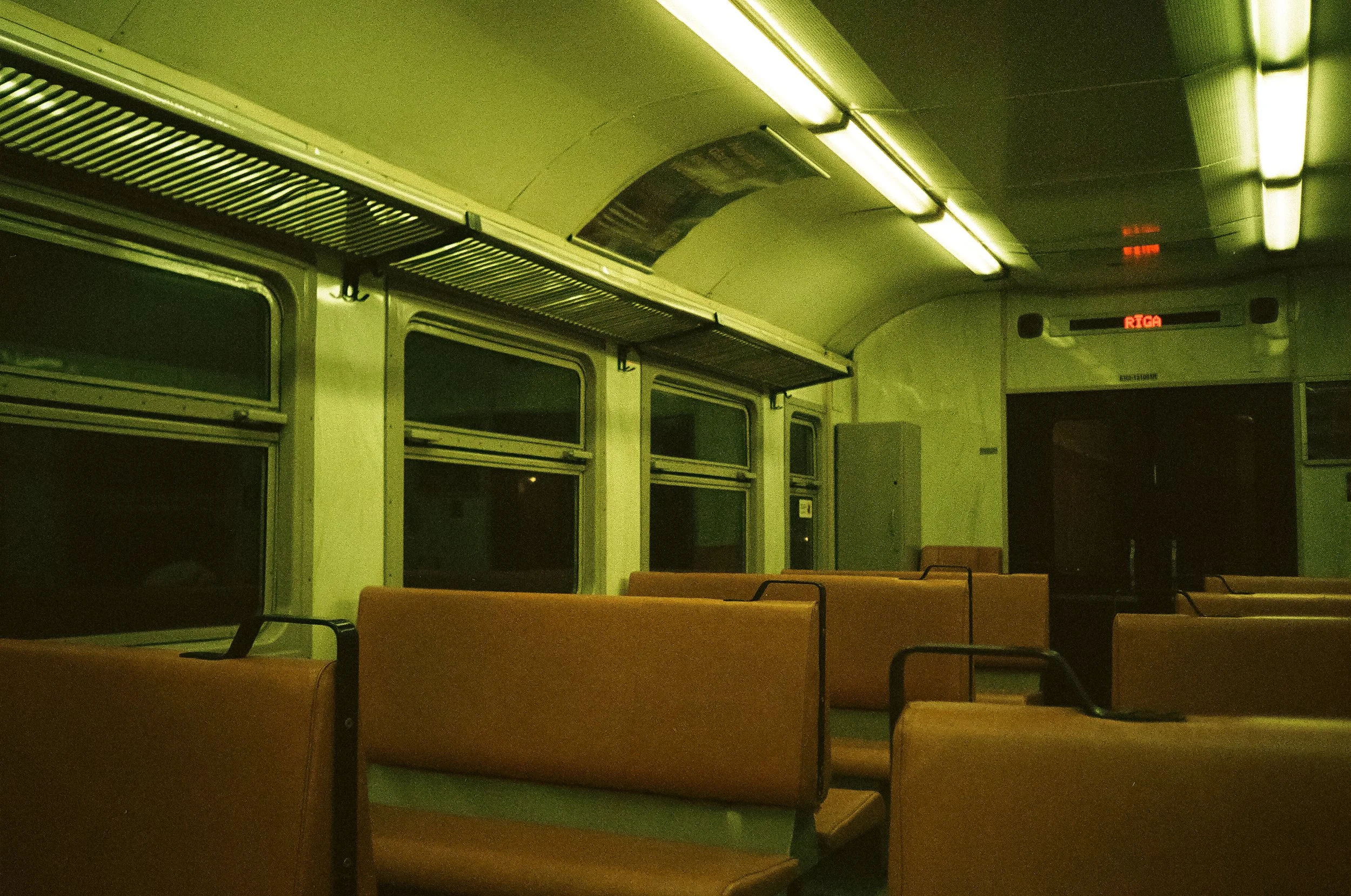 Interior of an empty train carriage at night, with rows of yellow seats, windows, luggage racks, fluorescent lighting, and a sign indicating RIGA at the front.