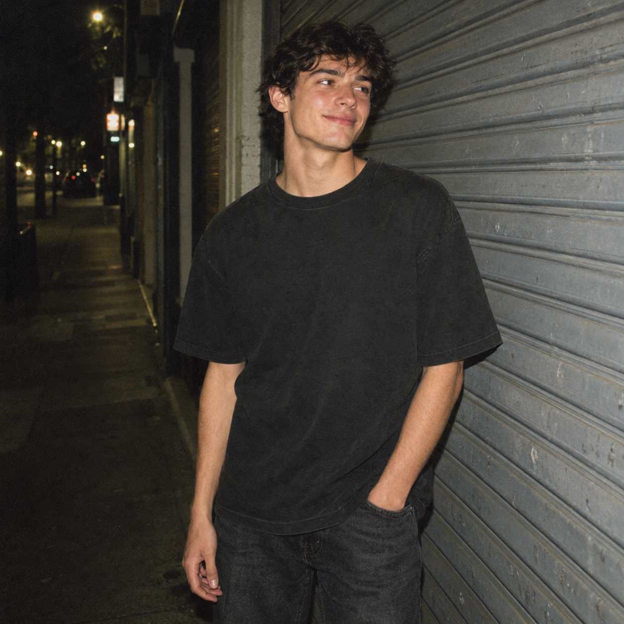A young man with curly dark hair and light skin standing on a sidewalk at night, wearing a black t-shirt and dark jeans, leaning against a metal shutter with his hands in his pockets and smiling.