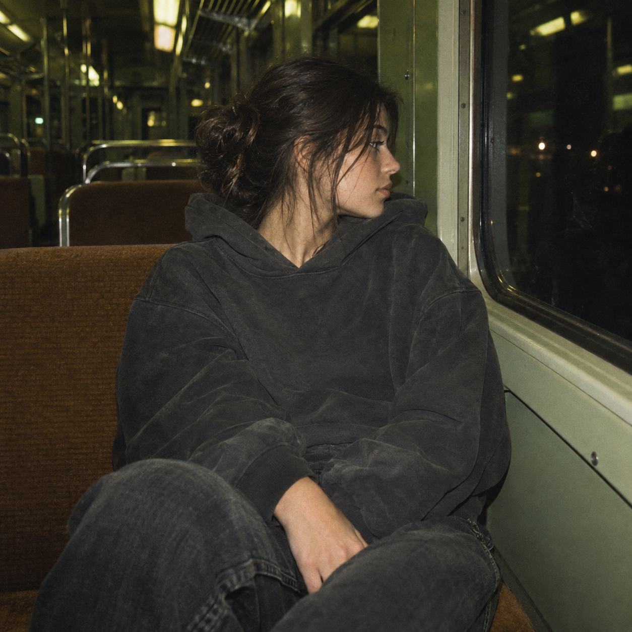 A young woman with dark hair tied in a messy bun, wearing a black hoodie and jeans, sitting alone on a bus at night, looking out the window with a contemplative expression.
