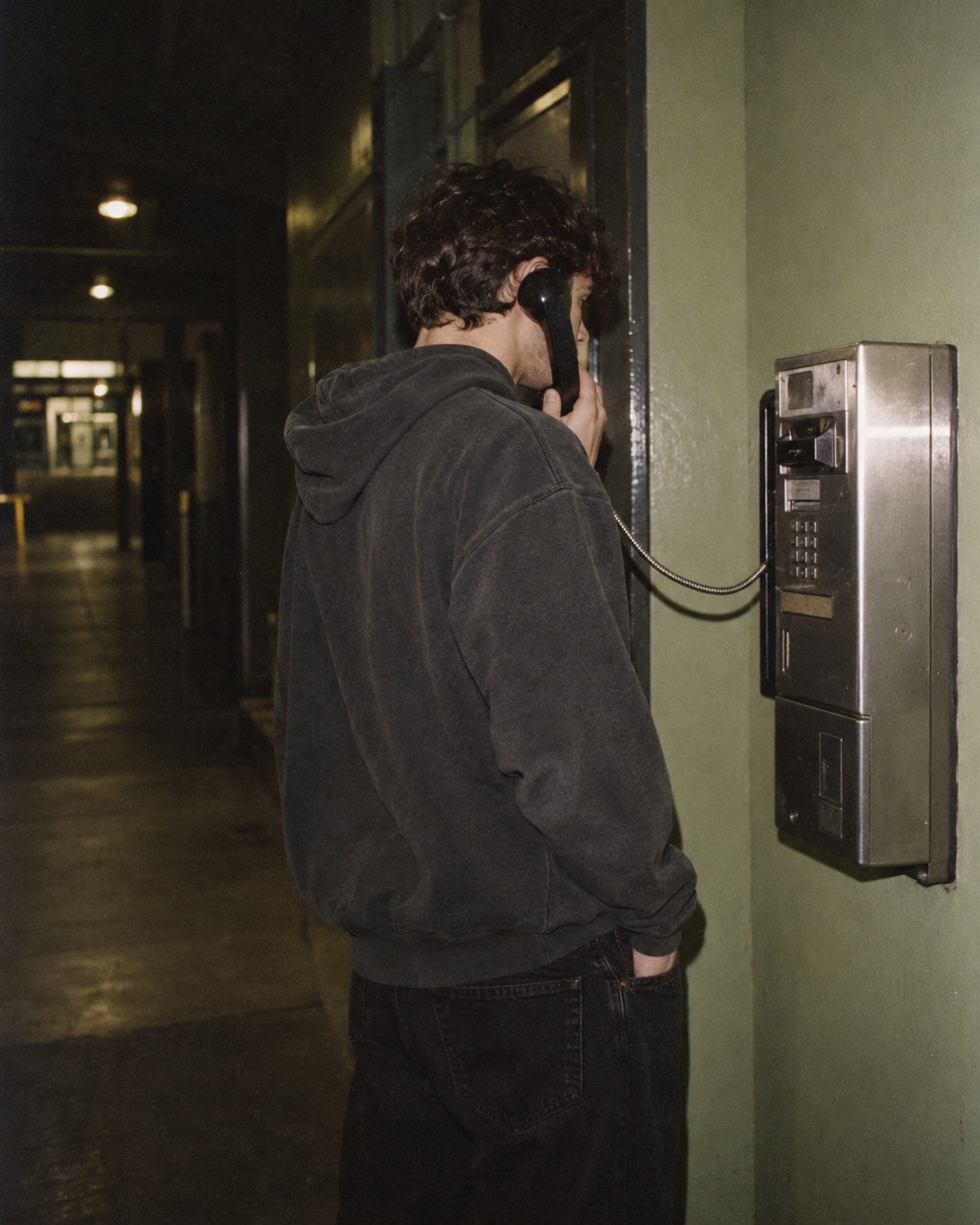 A young man in a black hoodie talking on a payphone in a hallway.