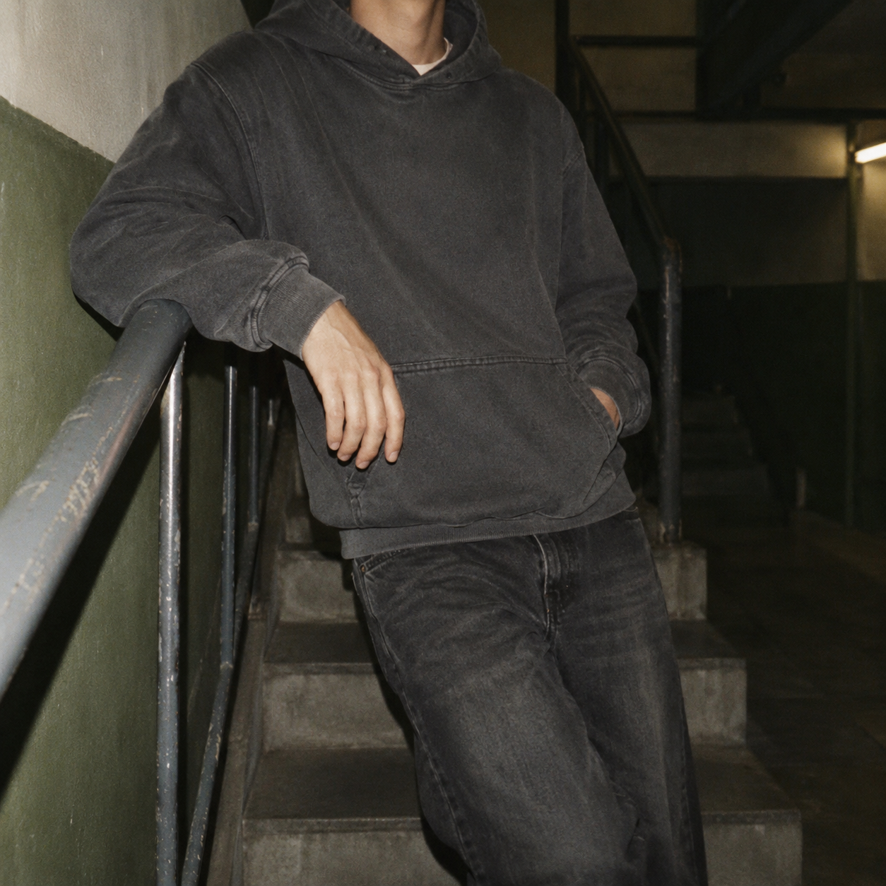 Person leaning against railing on a staircase, wearing a dark hoodie and jeans, in an industrial setting.