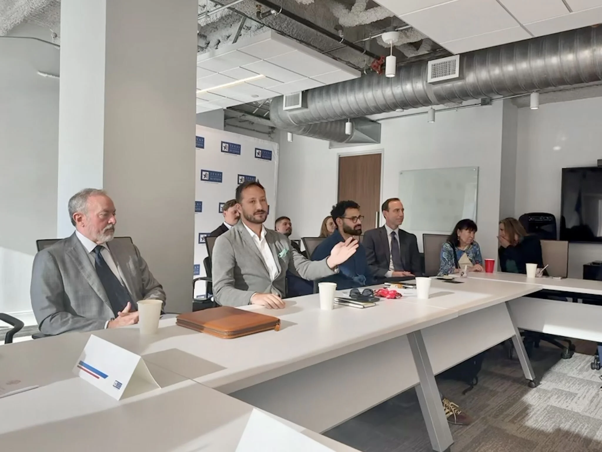 People attending a business meeting or conference in a modern office, sitting around a large white table, some with notebooks and cups, with exposed ceiling ducts and a whiteboard in the background.