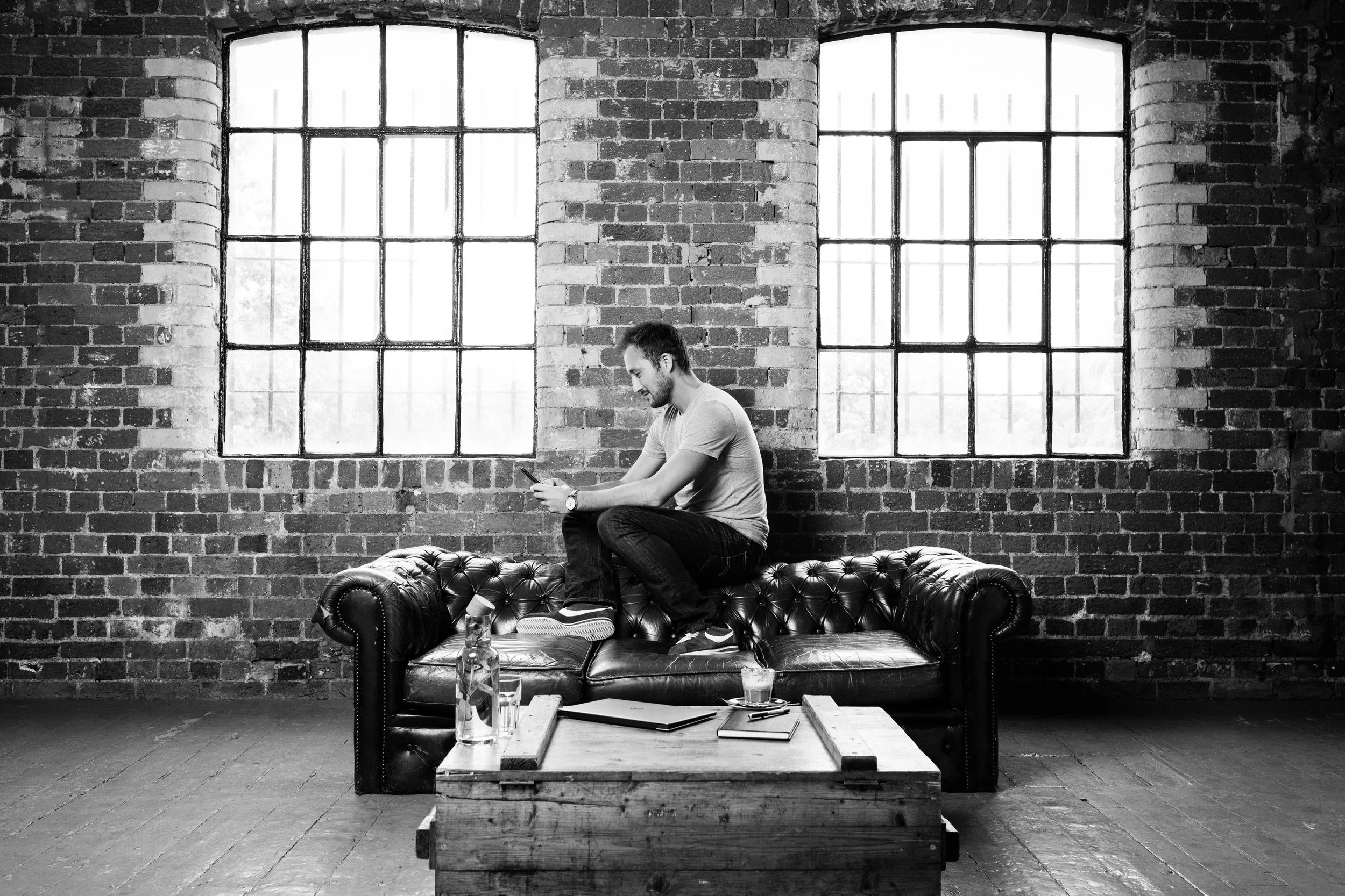 Joe Blackman sitting on a leather sofa in front of a brick wall, looking at his phone. There are large windows behind him with natural light coming in. A coffee table in front has a water bottle, glasses, notebooks, and a pen.