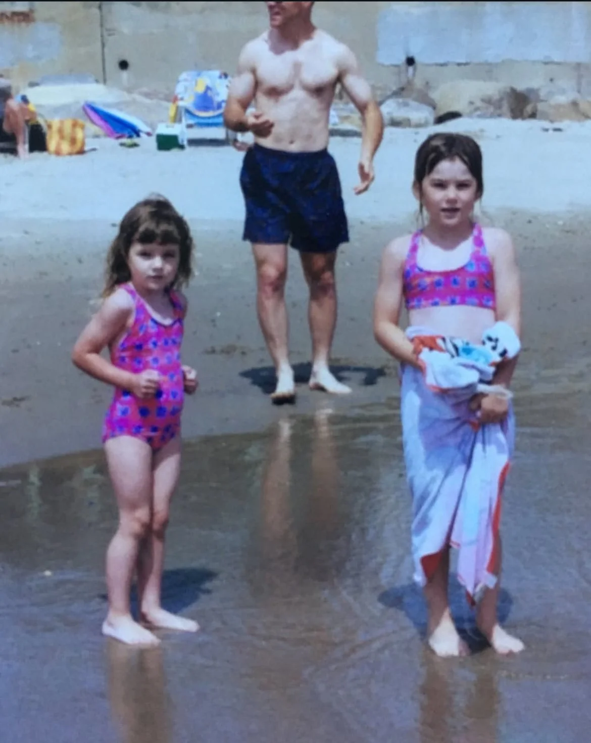 Two young girls in swimsuits standing on the beach with an adult man in swim trunks behind them, near the water.