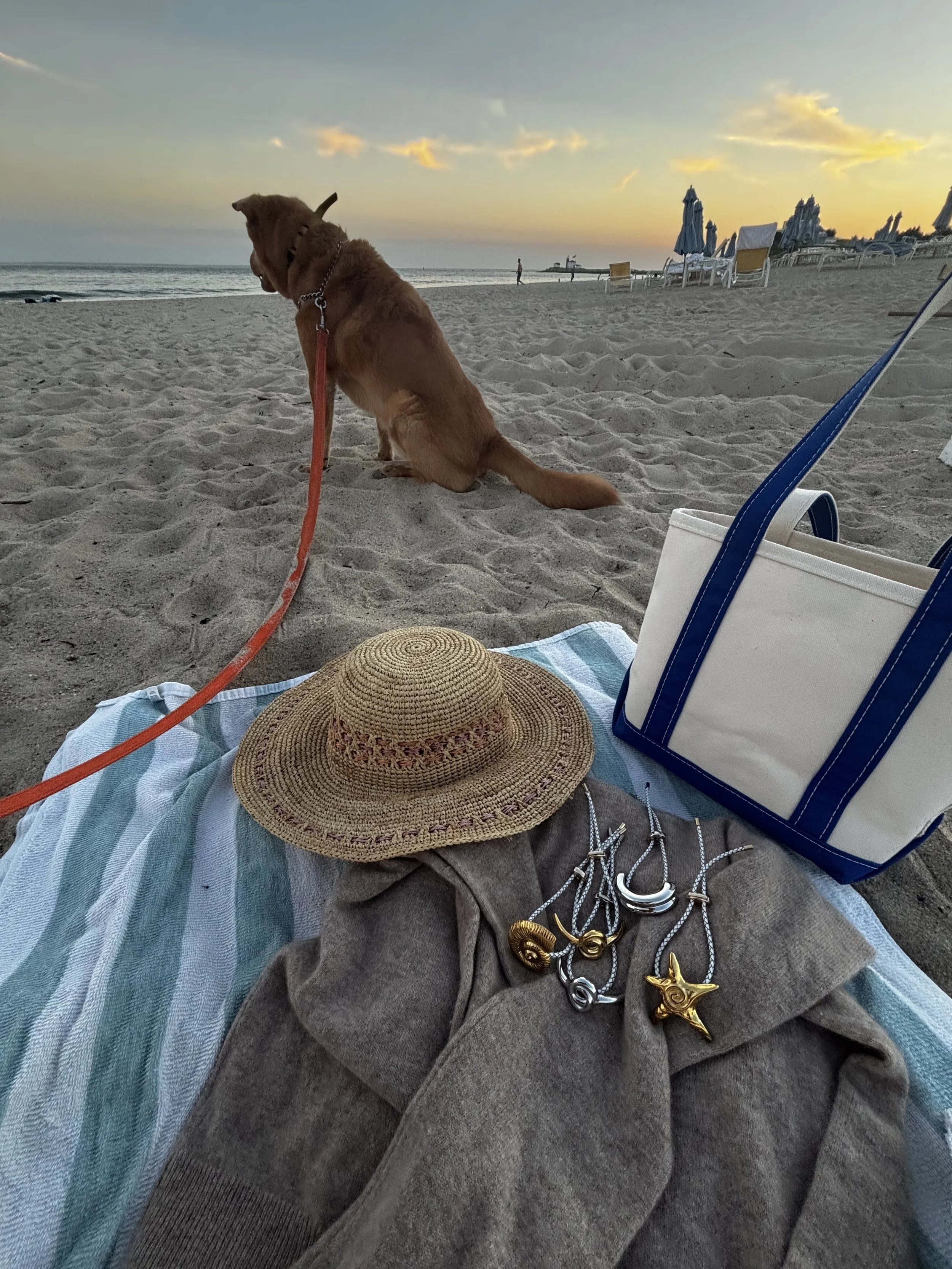 A sandy beach at sunset with lounge chairs and umbrellas in the background. In the foreground, a person is lying on a striped towel wearing jewelry, including necklaces with large pendant shells. A wide-brimmed straw hat and tote bag are also on the towel. A brown dog is sitting on the sand facing the water, with a leash attached to its collar.