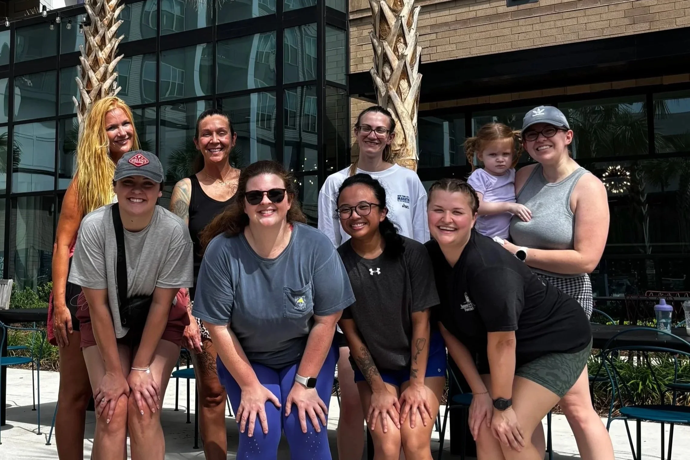 A group of nine women and girls pose together outdoors in front of a modern building with large glass windows and palm trees. They are smiling, standing close, some with hands on knees, and wearing casual athletic clothing.