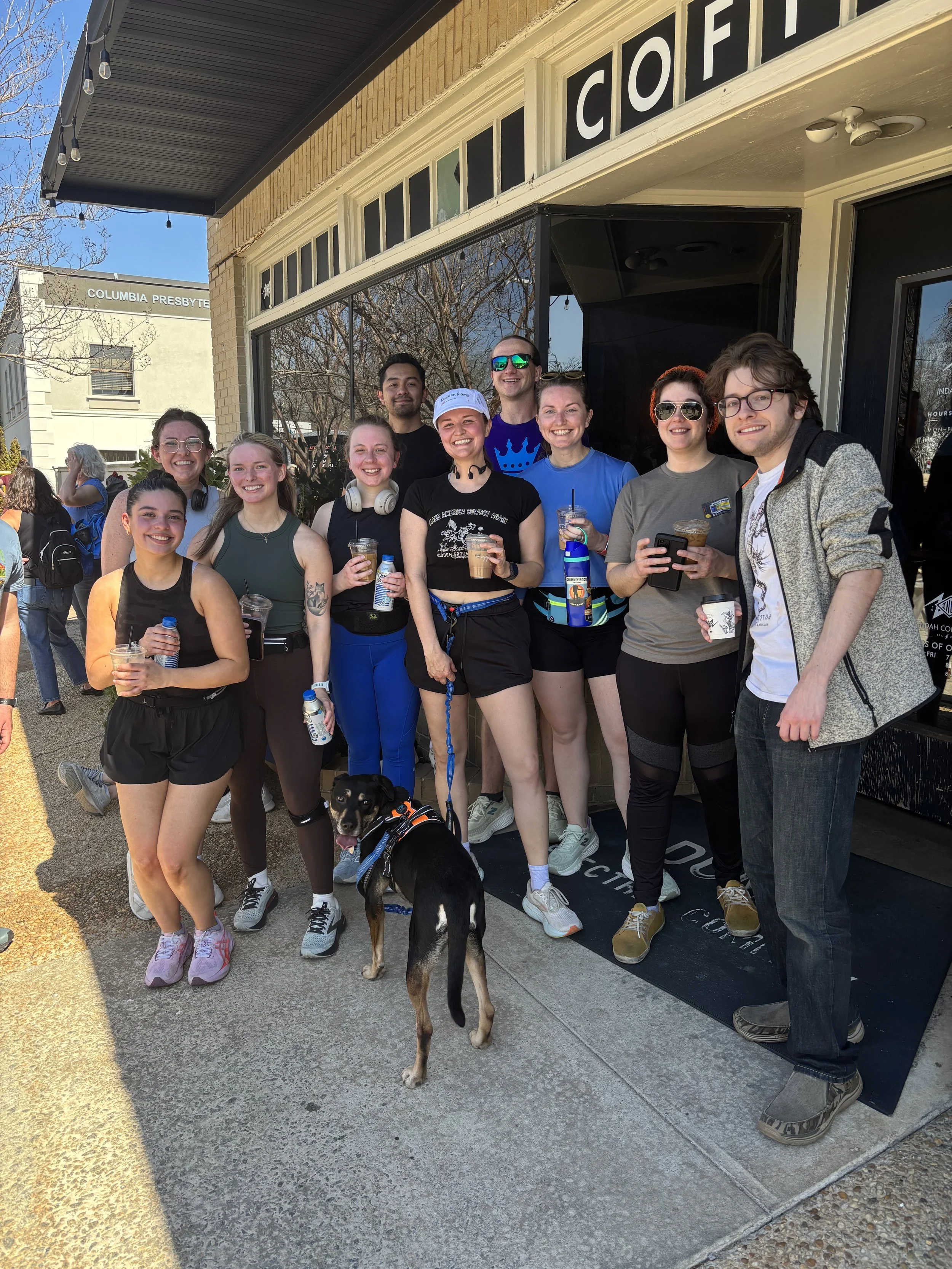 A group of ten people standing together outside a storefront with a dark door and window, smiling and holding drinks, along with a black dog with brown markings, in front of a sidewalk in daytime.
