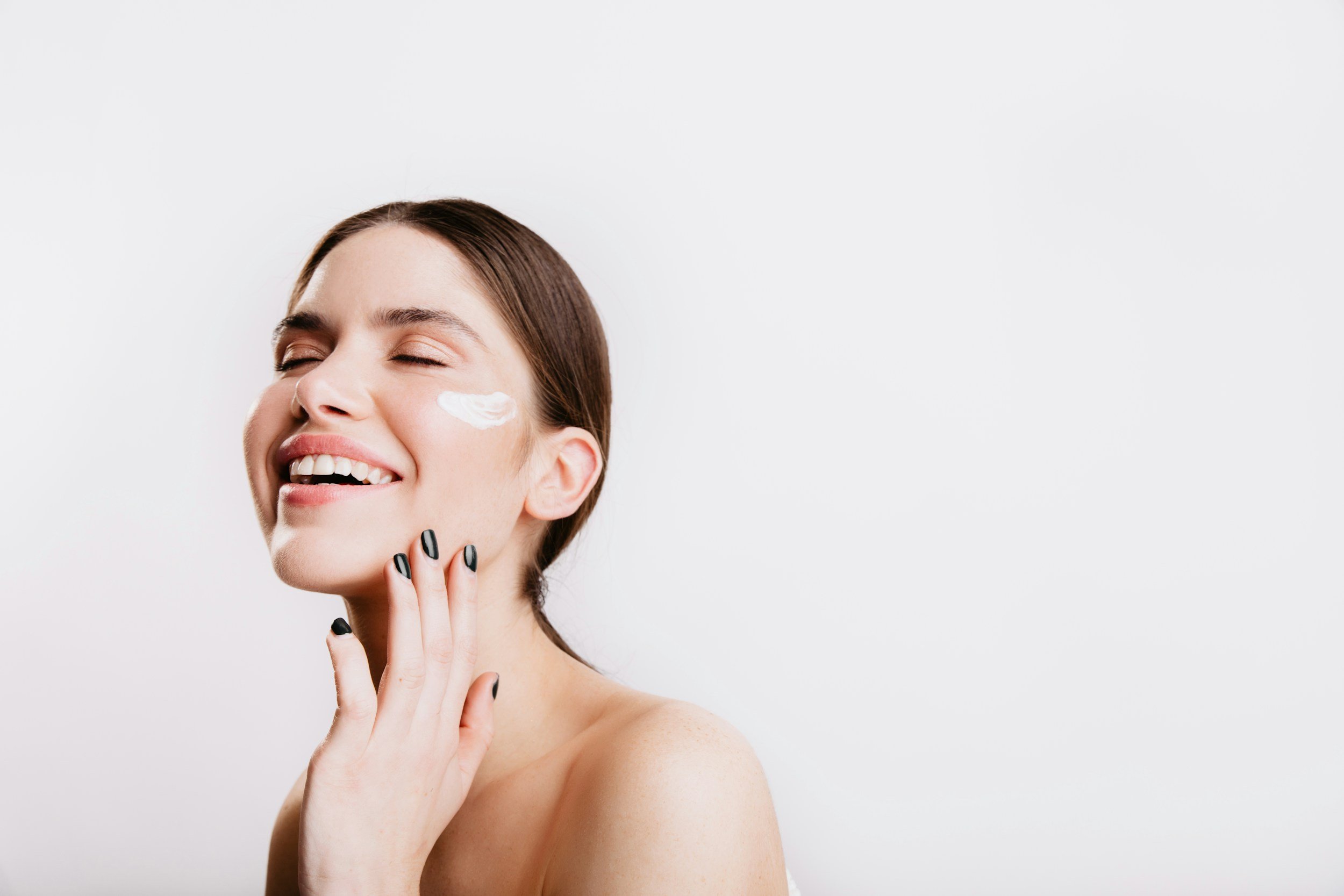 A woman smiling with her eyes closed, applying a white cream to her cheek, with her hand touching her face and black nail polish, against a plain white background.