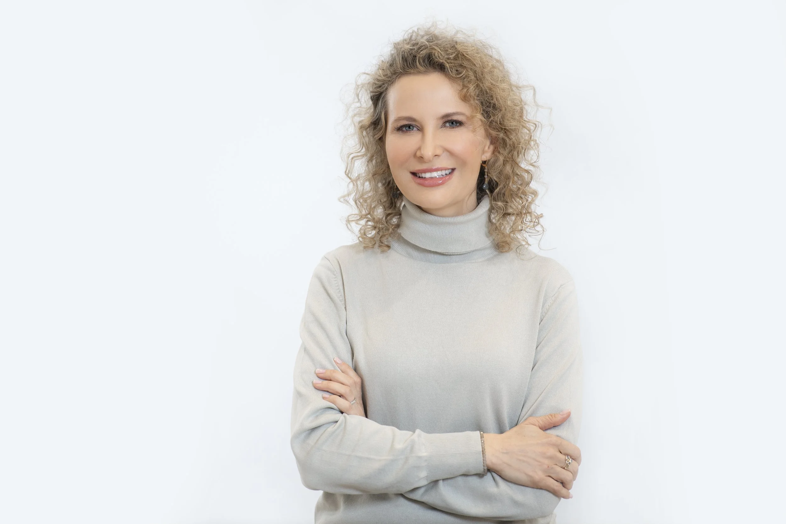A woman with curly blonde hair wearing a light-colored turtleneck sweater, smiling at the camera with her arms crossed in front of a plain white background.
