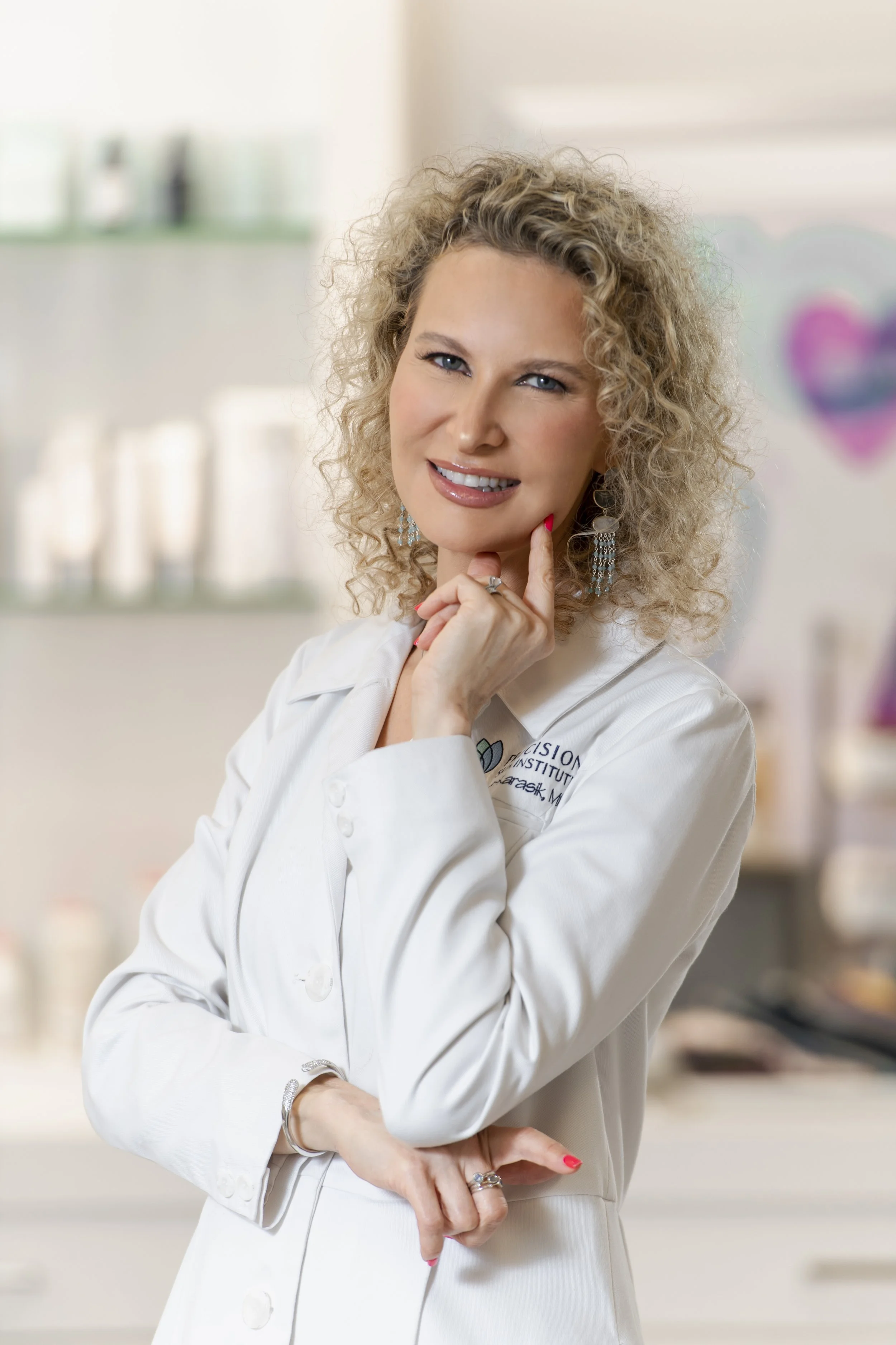 A woman in a white lab coat with curly hair and blue eyes, smiling and posing with her hand near her face, in a medical or clinical setting.