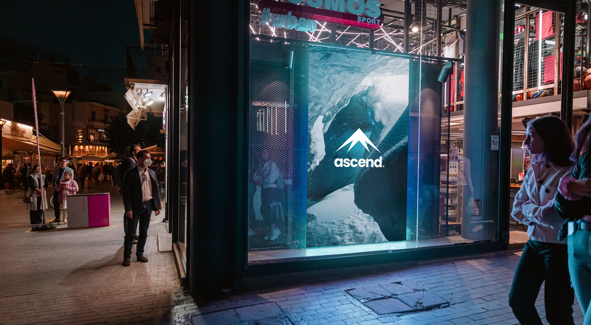 Nighttime street scene outside a Cosmos Sport store with an illuminated Ascend mountain advertisement in the window and pedestrians nearby.