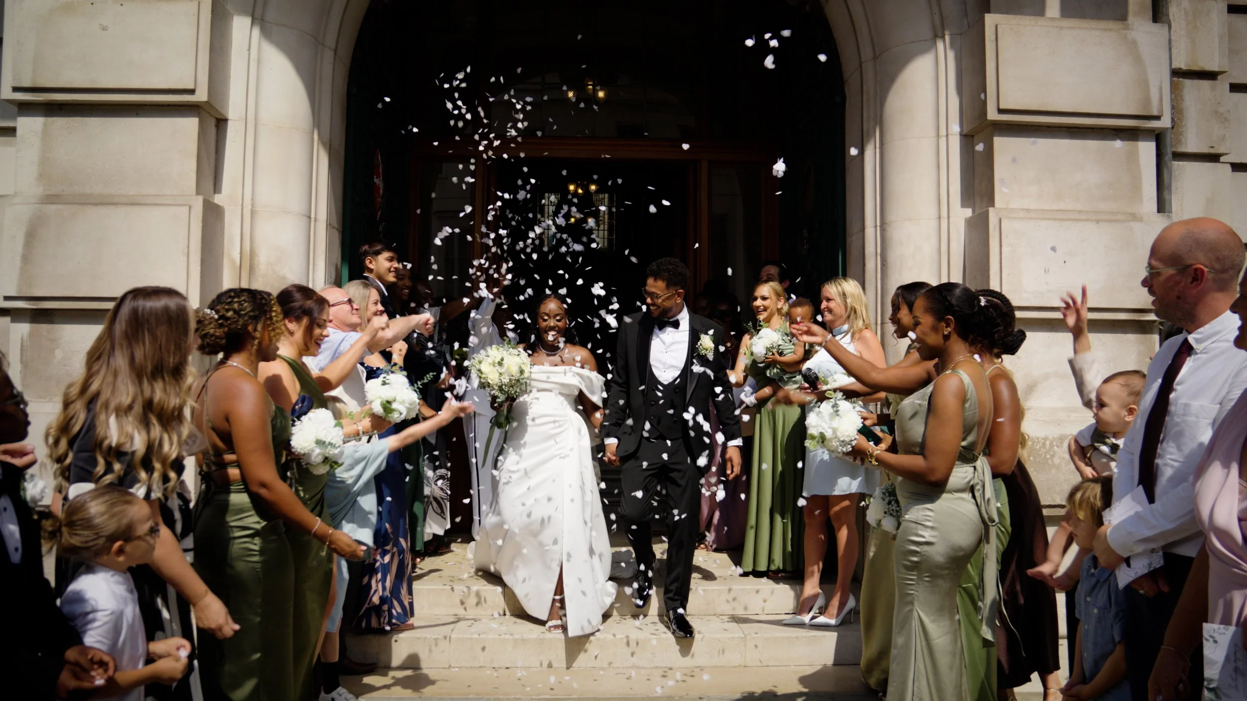 A wedding celebration with diverse guests throwing white flower petals as the newlywed couple steps out of a building with stone steps and a large arched entrance.