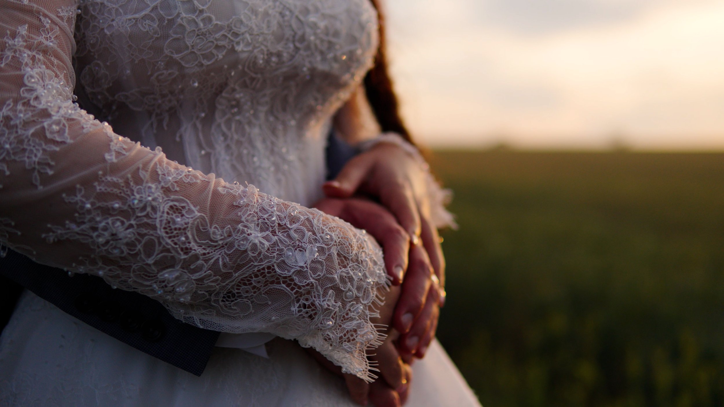 Close-up of a bride in a lace wedding dress holding hands with her groom, taken during sunset outdoors.