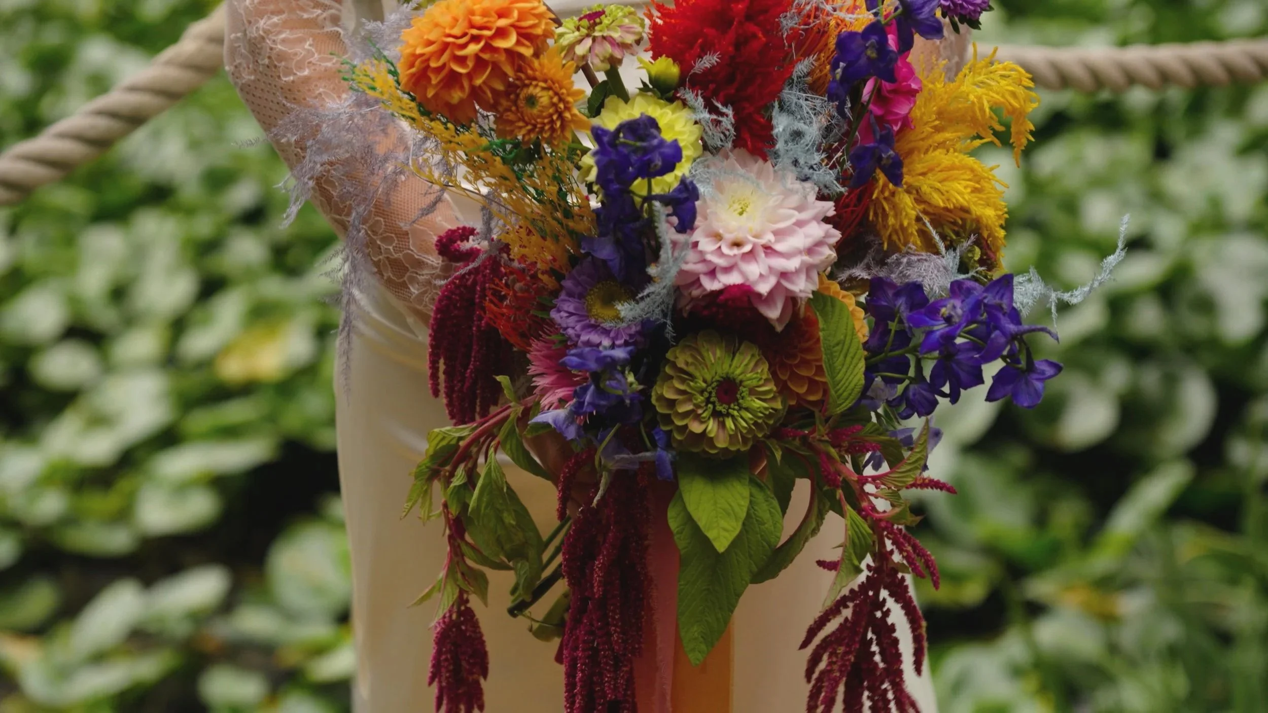 A person holding a colorful bouquet of various flowers, including pink, orange, yellow, purple, and red flowers with green leaves, wrapped in lace and fabric, in an outdoor setting.