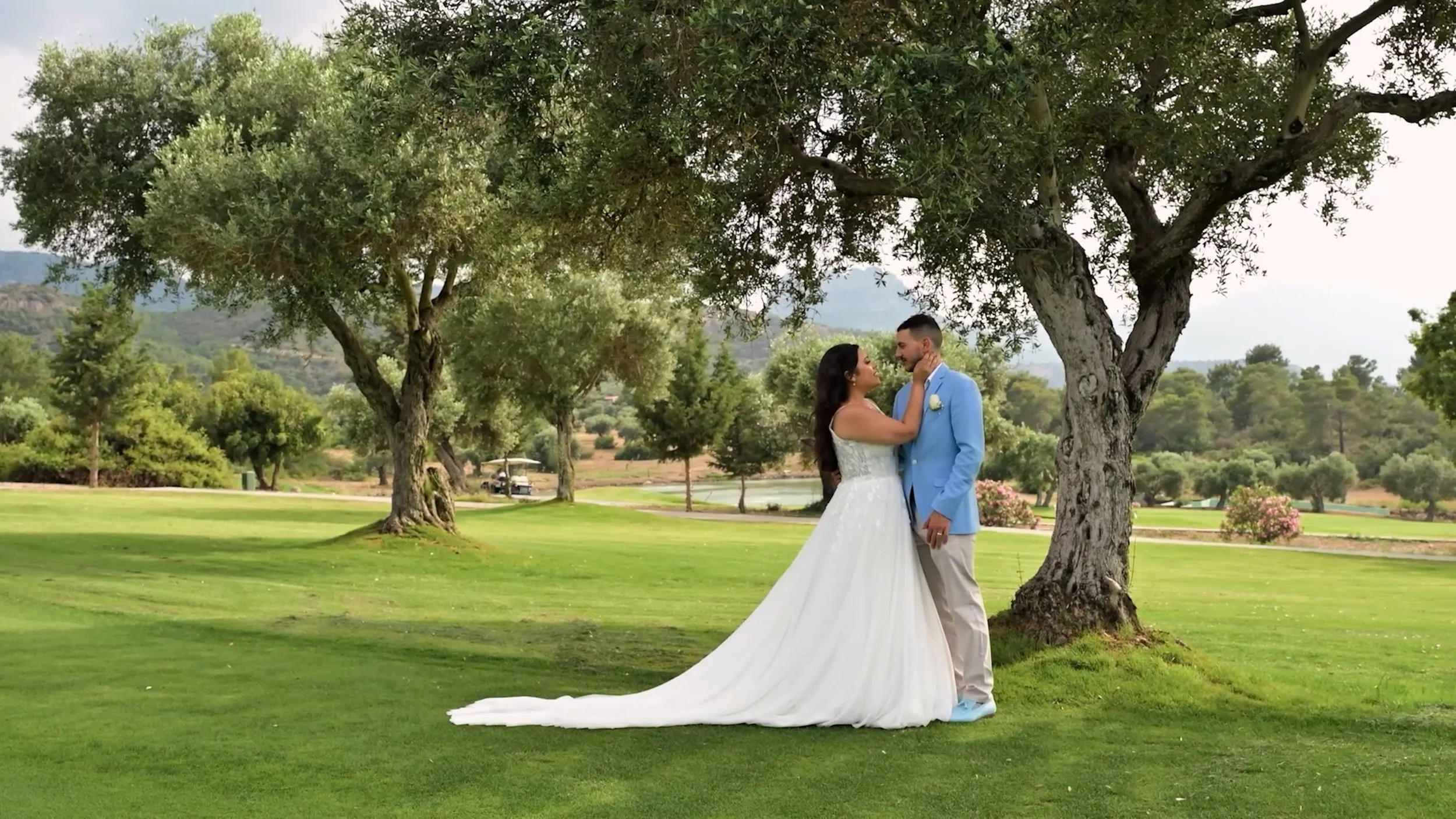 A bride and groom standing close together under a large tree on a golf course, with green grass and distant hills in the background, during daytime.