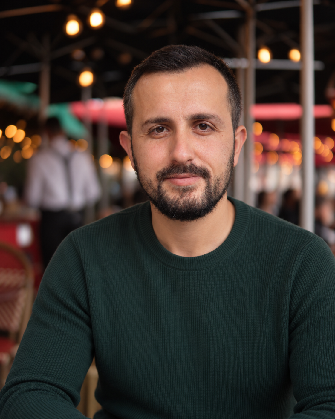 A man with short dark hair and a beard, wearing a dark green sweater, sitting indoors with blurred warm lighting and people in the background.