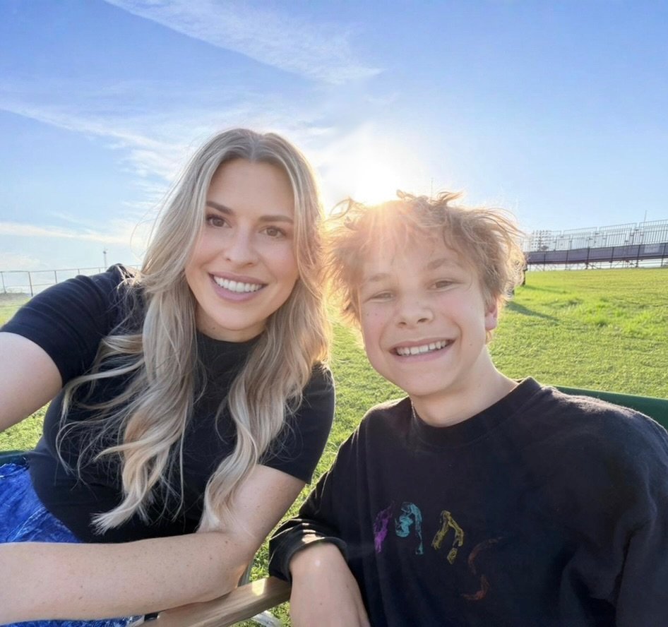 A woman and a boy smiling outdoors on a sunny day, sitting on some green chairs on a grassy field with a clear blue sky and a fence in the background.
