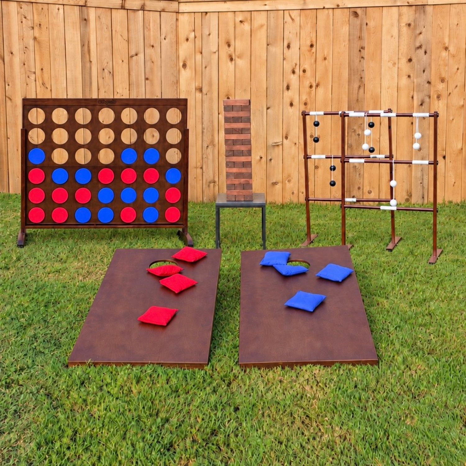 Outdoor scene with wooden board game, Jenga tower, connect four game, and tables with red and blue cushions, set on grass against a wooden fence.