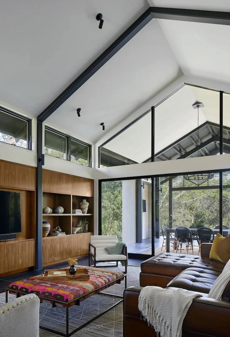 Modern living room with large windows, high ceiling, and a glass wall opening to an outdoor dining area, furnished with a leather sectional, a white armchair, and a colorful coffee table.