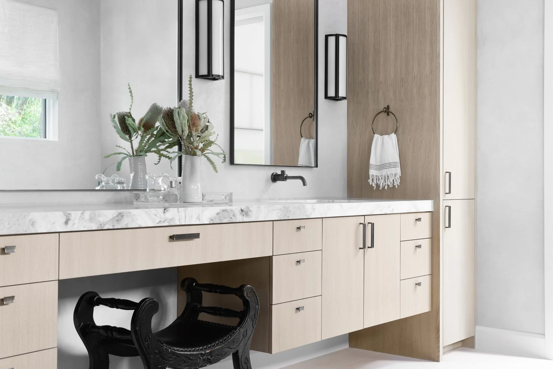A bathroom with a white marble countertop and wooden cabinets. A large mirror is mounted on the wall, with black wall sconces on either side. A small window with a white shade is on the left, and a towel ring with a white towel is on the right. There is a vase with green plants on the counter, and a black decorative chair is in front of the cabinetry.