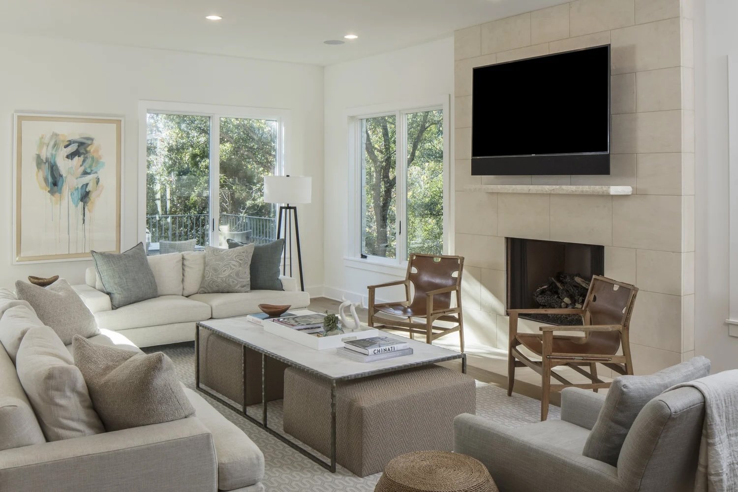 Living room with cream couches, a coffee table, two wooden chairs, a floor lamp, and large windows with trees outside. There's a wall-mounted flat-screen TV above a fireplace.