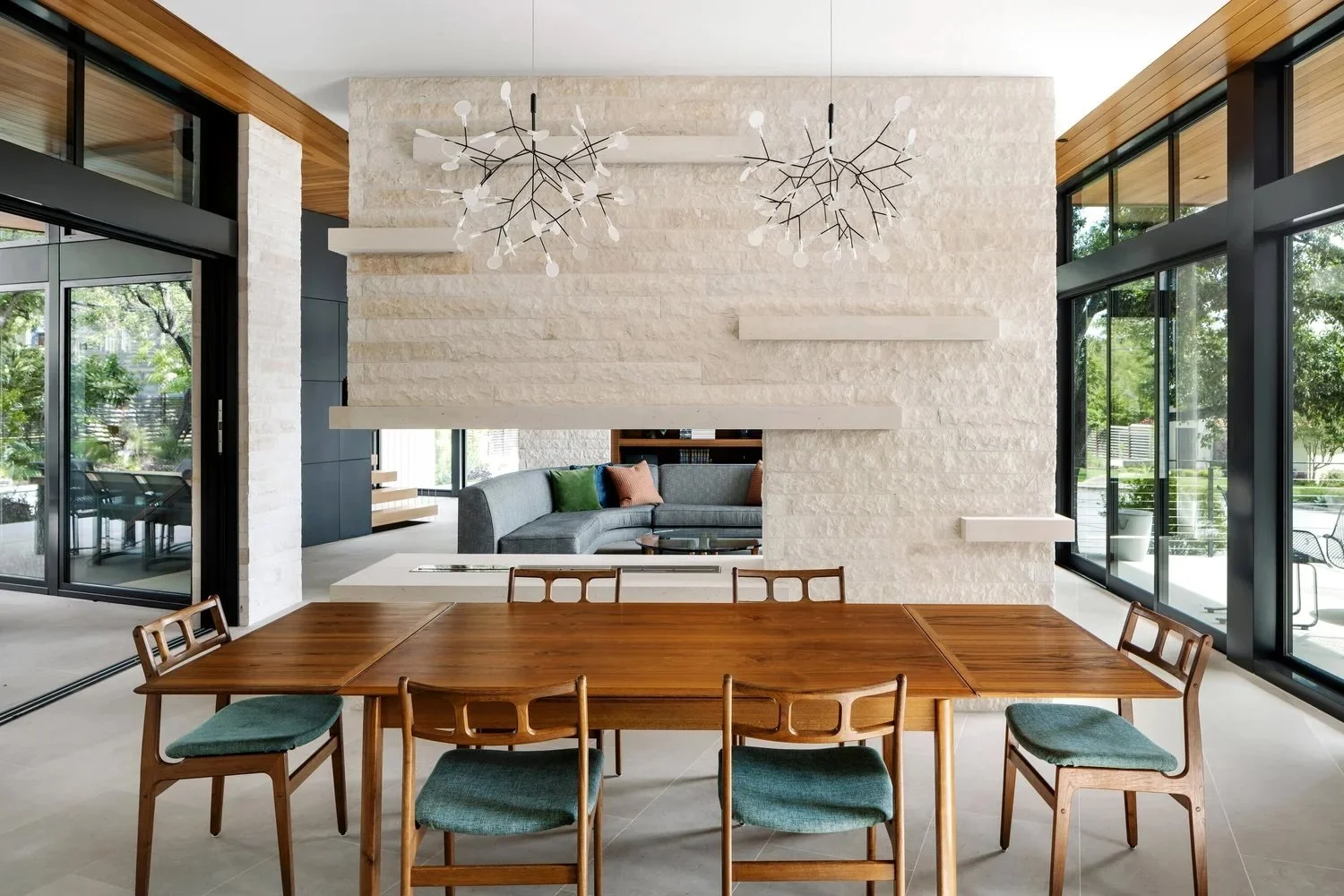 Modern dining room with wooden table and six chairs, large white brick wall with floating shelves, contemporary chandeliers, glass sliding doors leading to outdoor patio with trees and outdoor furniture.