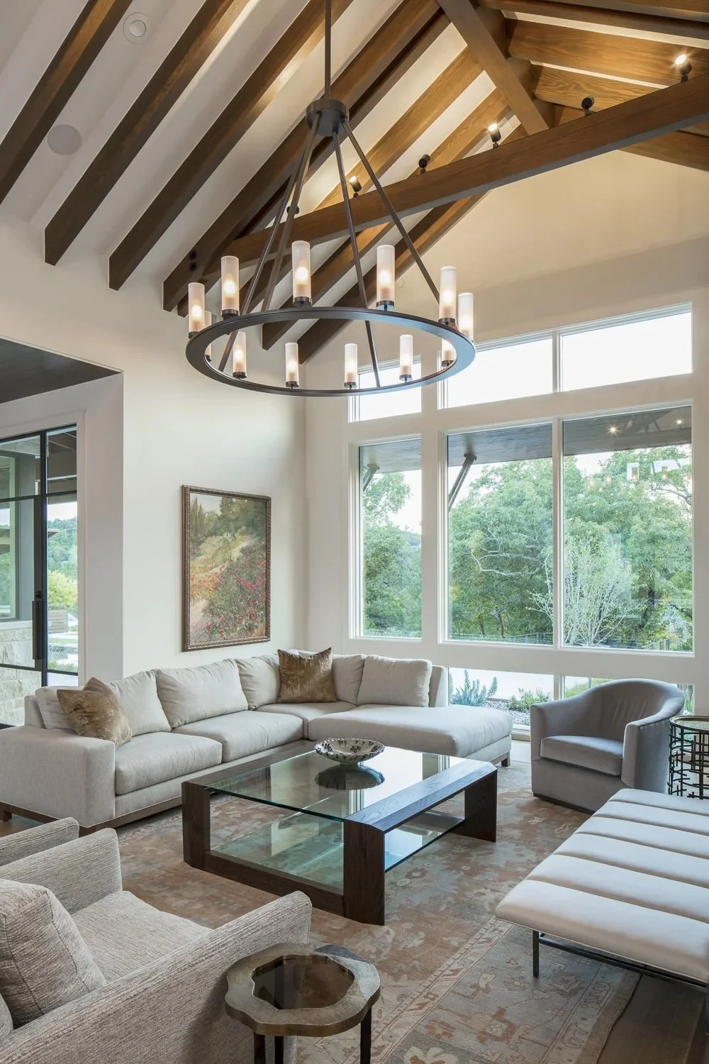 Living room with white sofa, beige armchair, glass coffee table, large windows showing green trees, wooden vaulted ceiling with beams, circular chandelier with candles.
