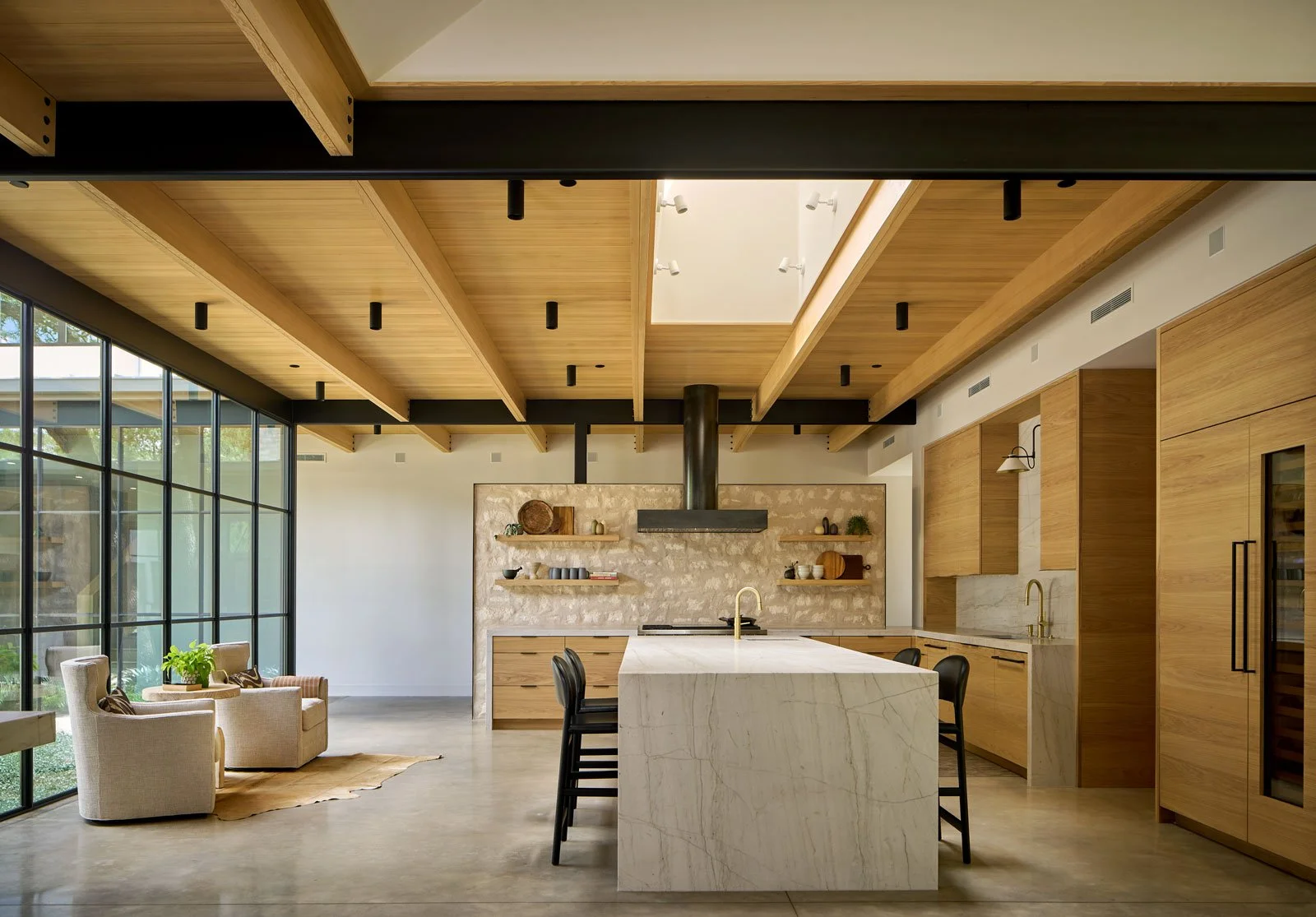 Modern kitchen with wooden cabinets, marble island, and open shelving, sunlight streaming through large glass windows, beige armchairs with cushions in a cozy seating area.