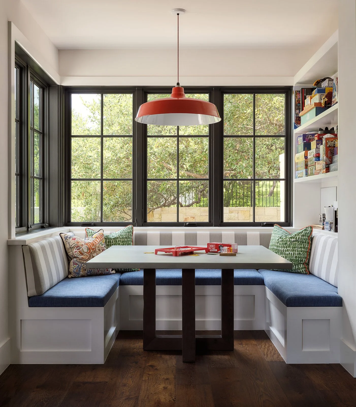 Kitchen nook with built-in white bench seating, green and patterned throw pillows, a white table, and large black-framed windows showing greenery outside. Red pendant light hangs above the table, and a shelf with books is on the right.