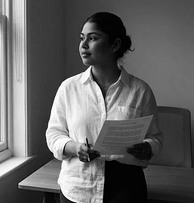 A woman with dark hair pulled back stands near a window, holding a paper and a pen, looking out the window in an indoor setting.