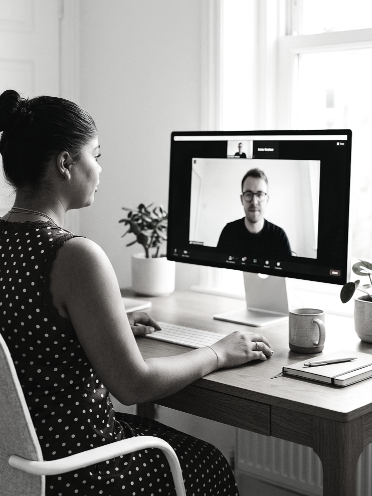 A woman with dark hair tied in a bun, wearing a polka dot sleeveless top, sitting at a desk and participating in a video call on a computer. The screen shows a man with glasses in a dark shirt. The desk has a notebook, pen, mug, and potted plants. There is a window with natural light in the background.