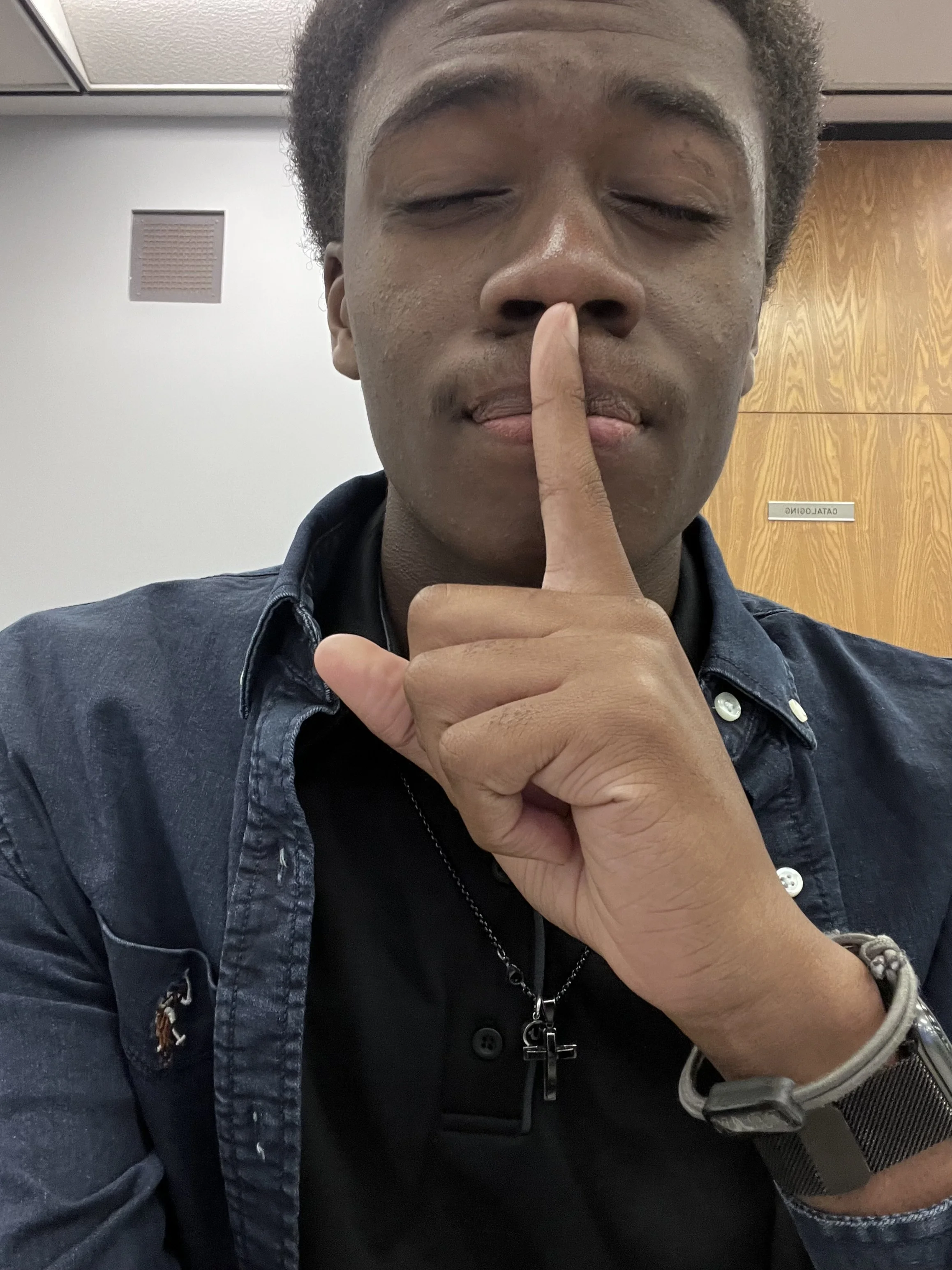 Close-up of a young man with curly hair, eyes closed, holding his finger to his lips in a 'shh' gesture, wearing a dark denim jacket and jewelry, in an indoor setting with wooden and white walls.