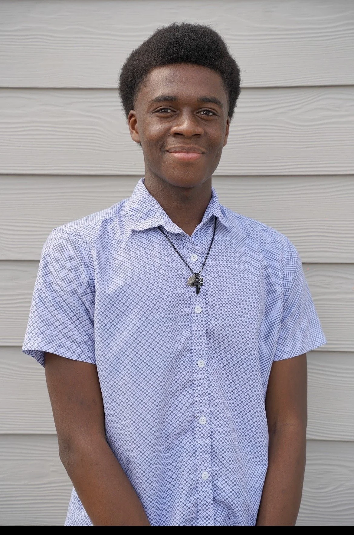 A young person with dark curly hair smiling, wearing a light blue patterned short-sleeve button-up shirt and a black necklace with a small pendant, standing in front of a light-colored horizontal siding background.