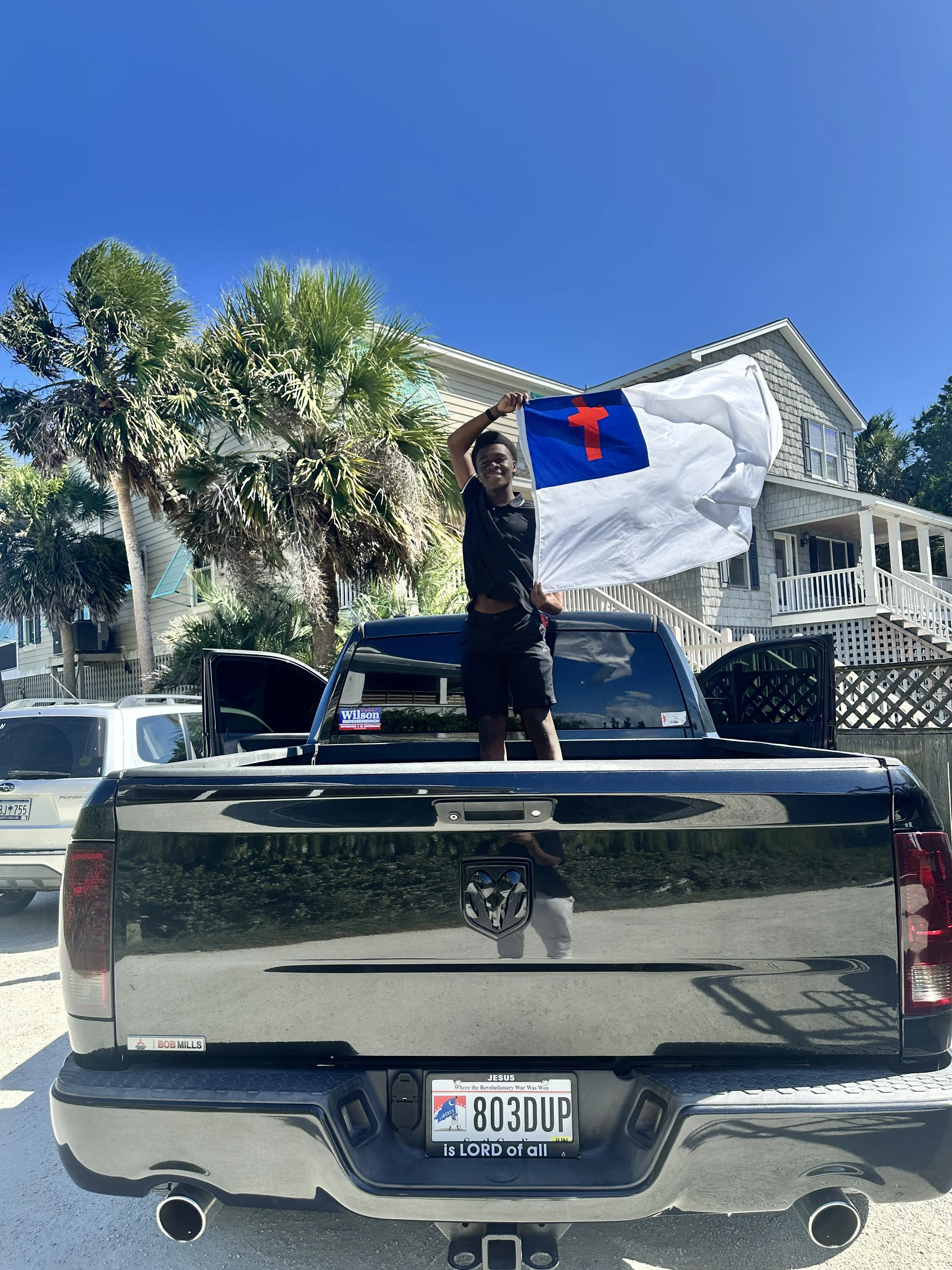Person standing in the truck bed holding a flag with a blue field and red cross. The truck has a shiny black rear with a Jesus sticker on the license plate frame and a bumper sticker that reads "Where the Revolutionary War Was Won." The background features palm trees, a residential house, and a clear blue sky.