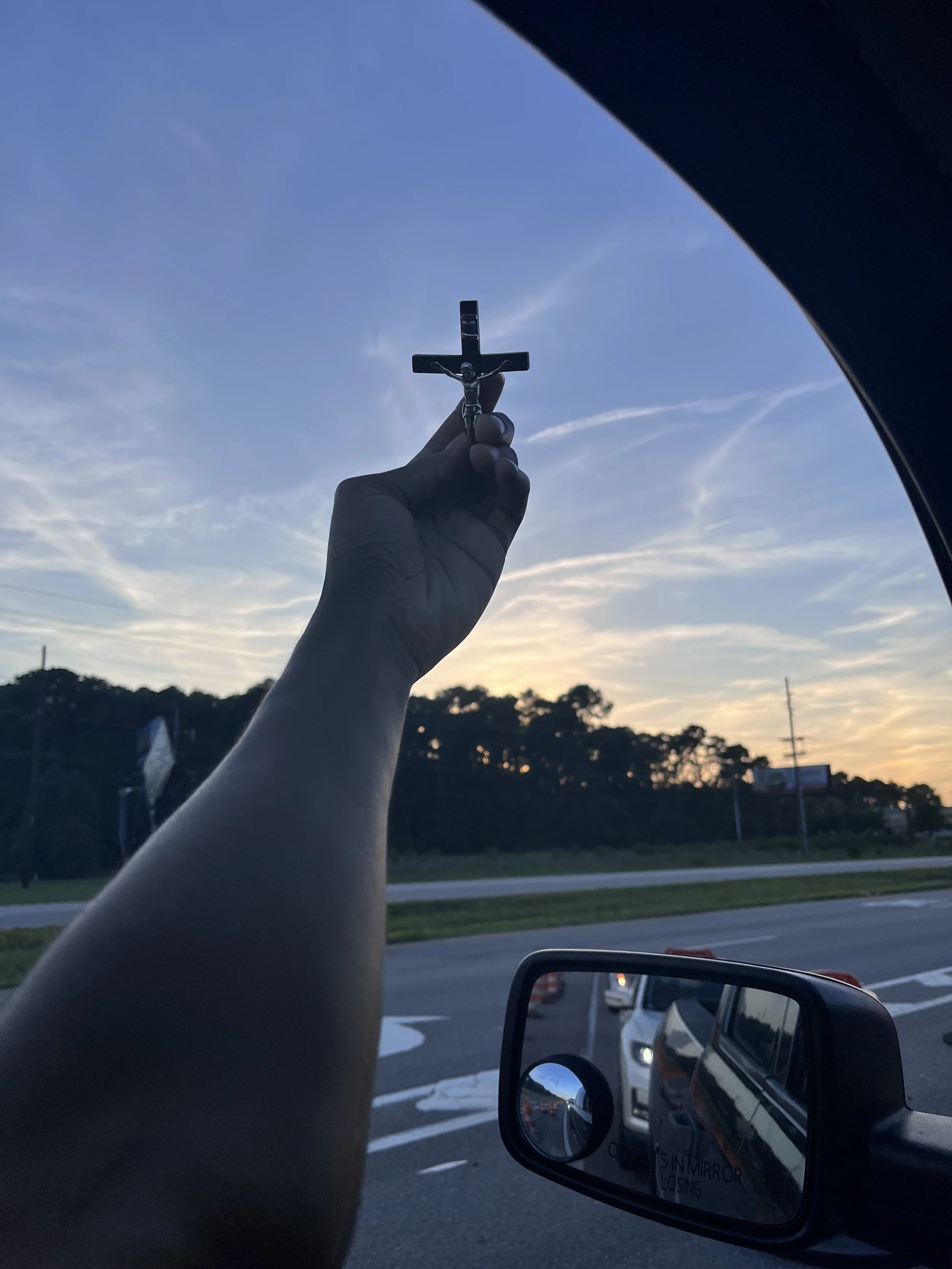 A hand holding a small crucifix with Jesus in front of a blue sky with some clouds, near sunset.