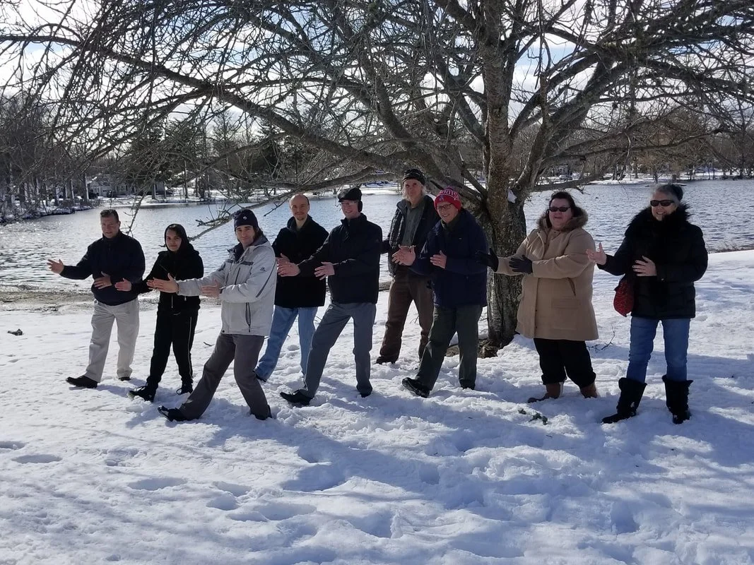 Group of nine adults standing in snow near a large leafless tree by a lake, posing with their hands extended forward.