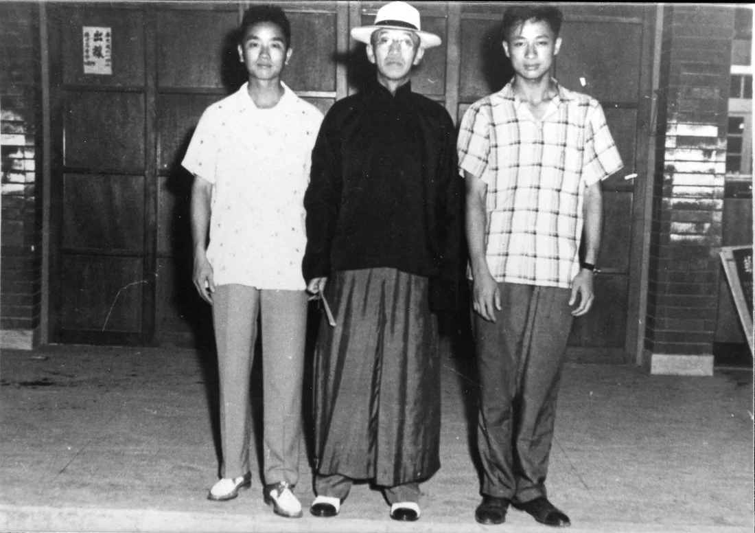Black and white photo of three people standing indoors in front of wooden panels. The person in the middle is wearing traditional Asian clothing and a wide-brimmed hat, holding a small object, flanked by two young men in casual Western attire, all looking at the camera.