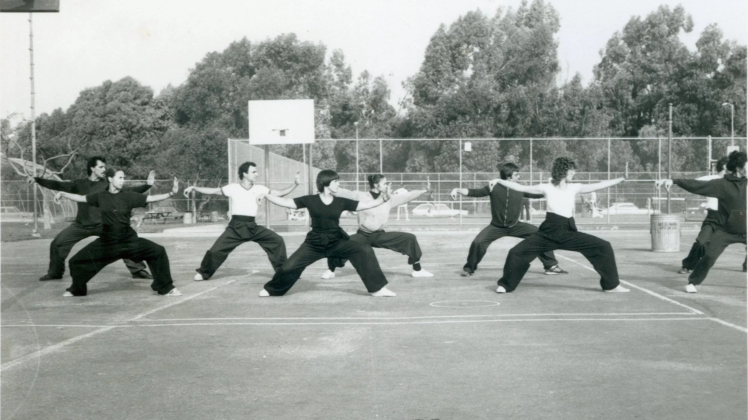 Group of people practicing martial arts on an outdoor basketball court, with trees and a fence in the background, in black and white.
