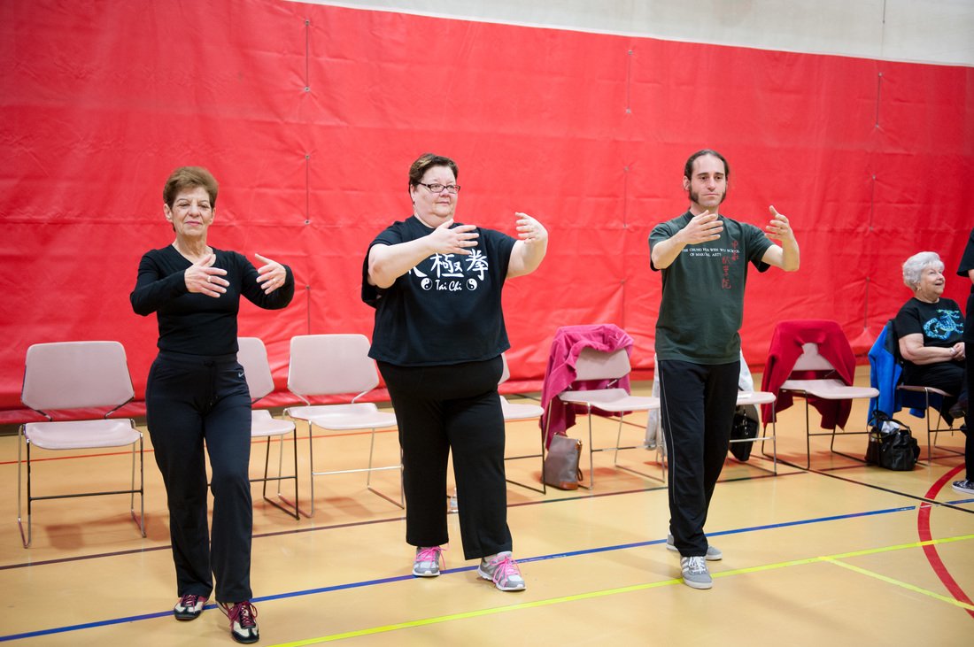 Three people practicing Tai Chi in a gym, standing with arms raised and bent at the elbows, with chairs and a red wall in the background.