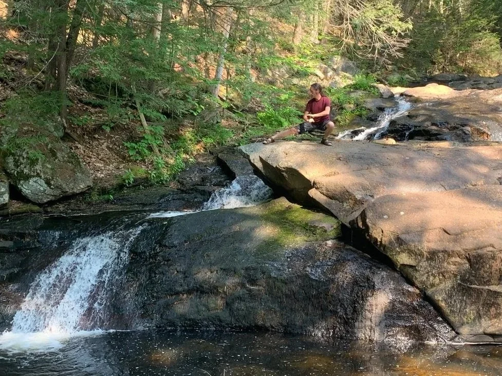 A person sitting on large rocks by a small waterfall in a forested area, surrounded by green trees and foliage.