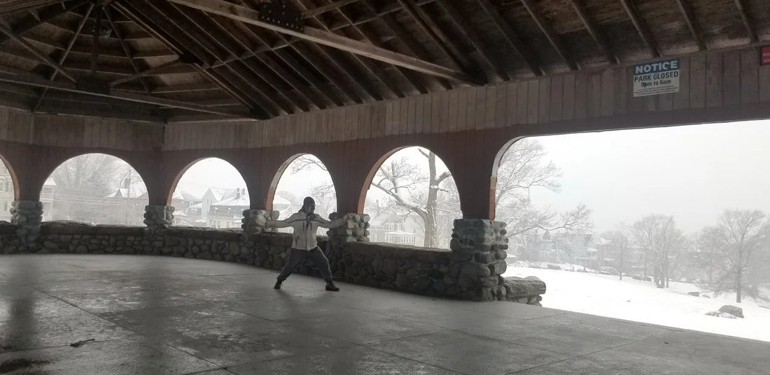 A person performing a martial arts move inside a covered outdoor space with stone and wood construction, overlooking a snowy landscape with trees and houses.