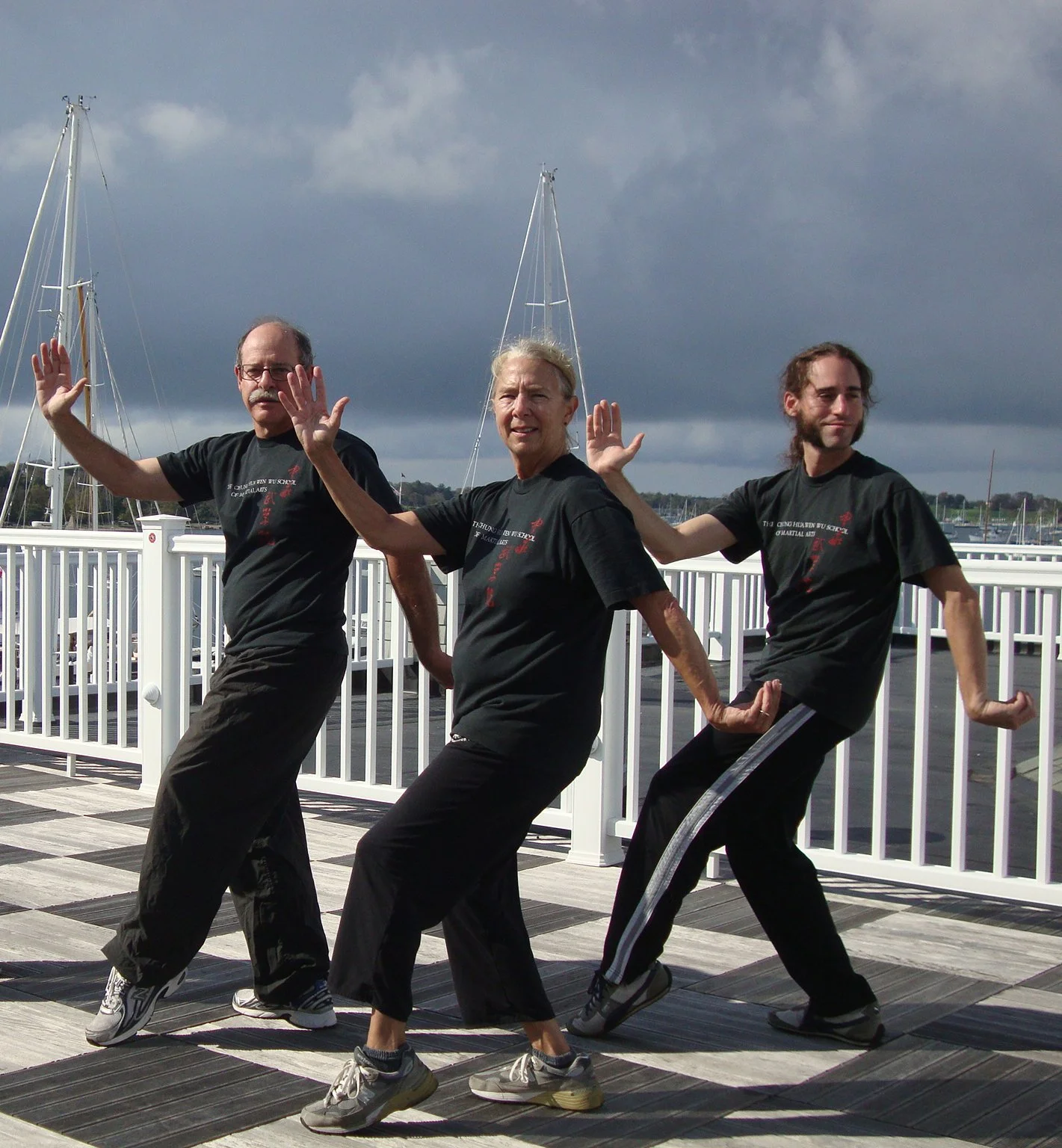 Three men practicing martial arts on a outdoor deck near the water, with sailboats and cloudy sky in the background.