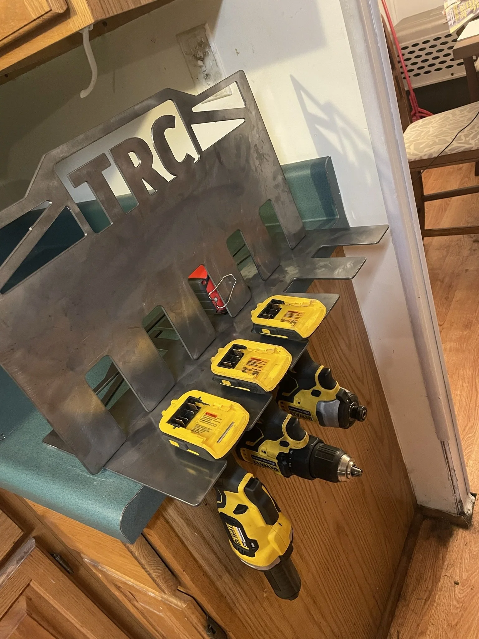 Tools on a counter including a cordless drill, yellow tool cases, and a metal 'TRICK' sign, with chairs and a table in the background.
