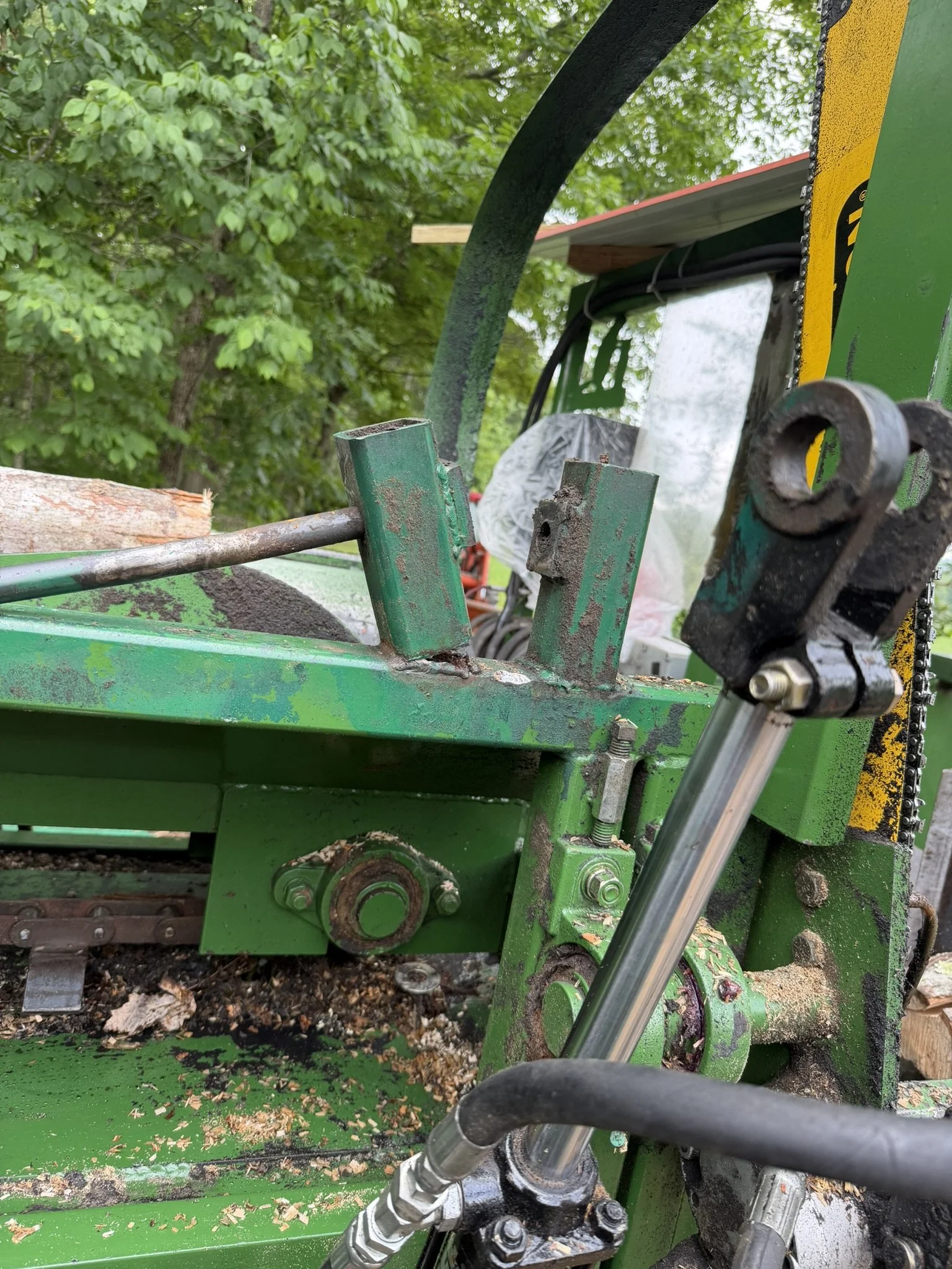 Close-up of green machinery with hydraulic components and wooden logs, with trees and greenery in the background.