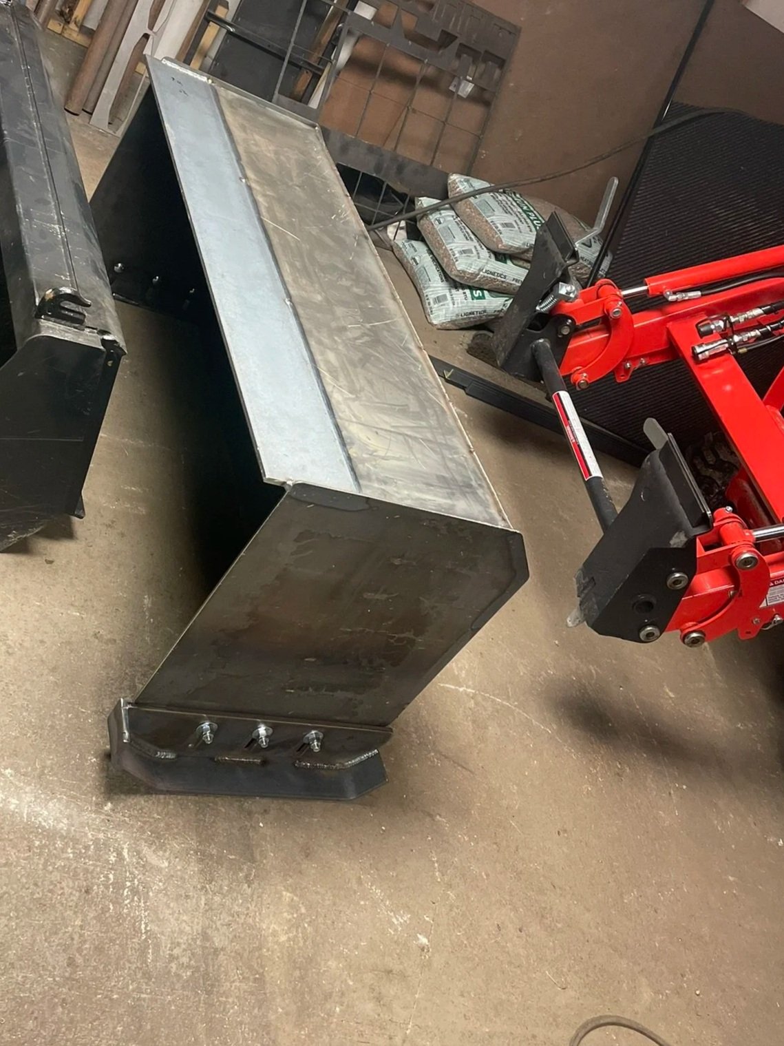 A metal workbench with a flat surface and a storage cabinet underneath, placed on a concrete floor in a workshop. Parts of a red hydraulic machine or lift are visible nearby, along with some pipes or other equipment in the background.