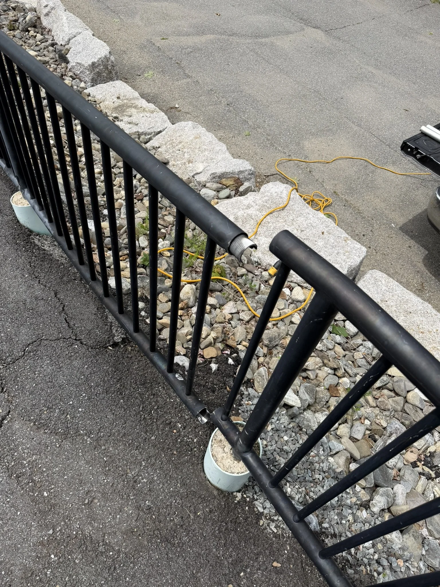 A black metal fence with vertical bars and white cylindrical bases, next to a gravel bed with small rocks and a concrete curb, separating it from an asphalt parking area with a power cord and a black toolbox.