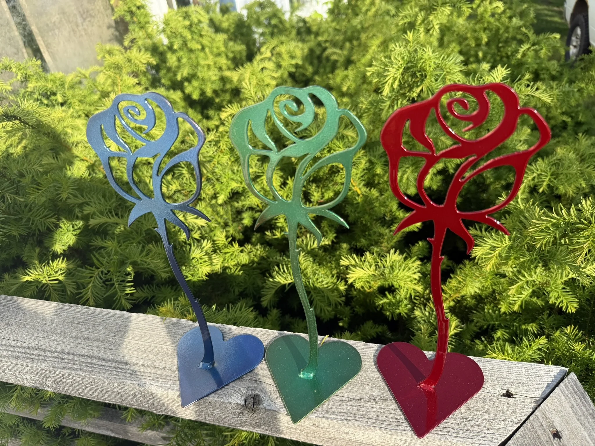 Three metal rose-shaped garden stakes in blue, green, and red, standing on a wooden surface with green foliage in the background.